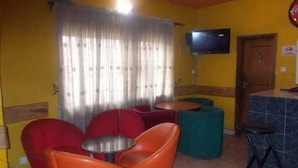 A room with yellow walls, a TV mounted on the wall, a wooden door, and a window with curtains. There are orange and red chairs, a small round table, and a teal ottoman. - Class Suites Signature (Lagos)