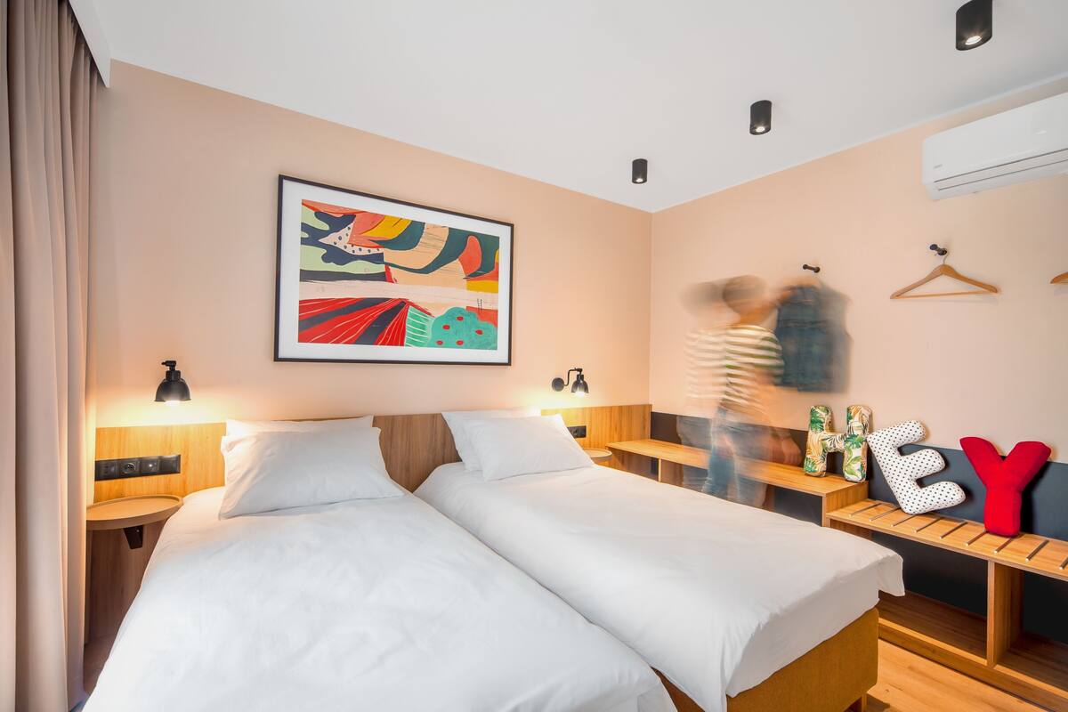 Ein Hotelzimmer mit zwei Betten, einem Nachttisch, einer Wand-Klimaanlage und einem farbenfrohen, abstrakten Gemälde.