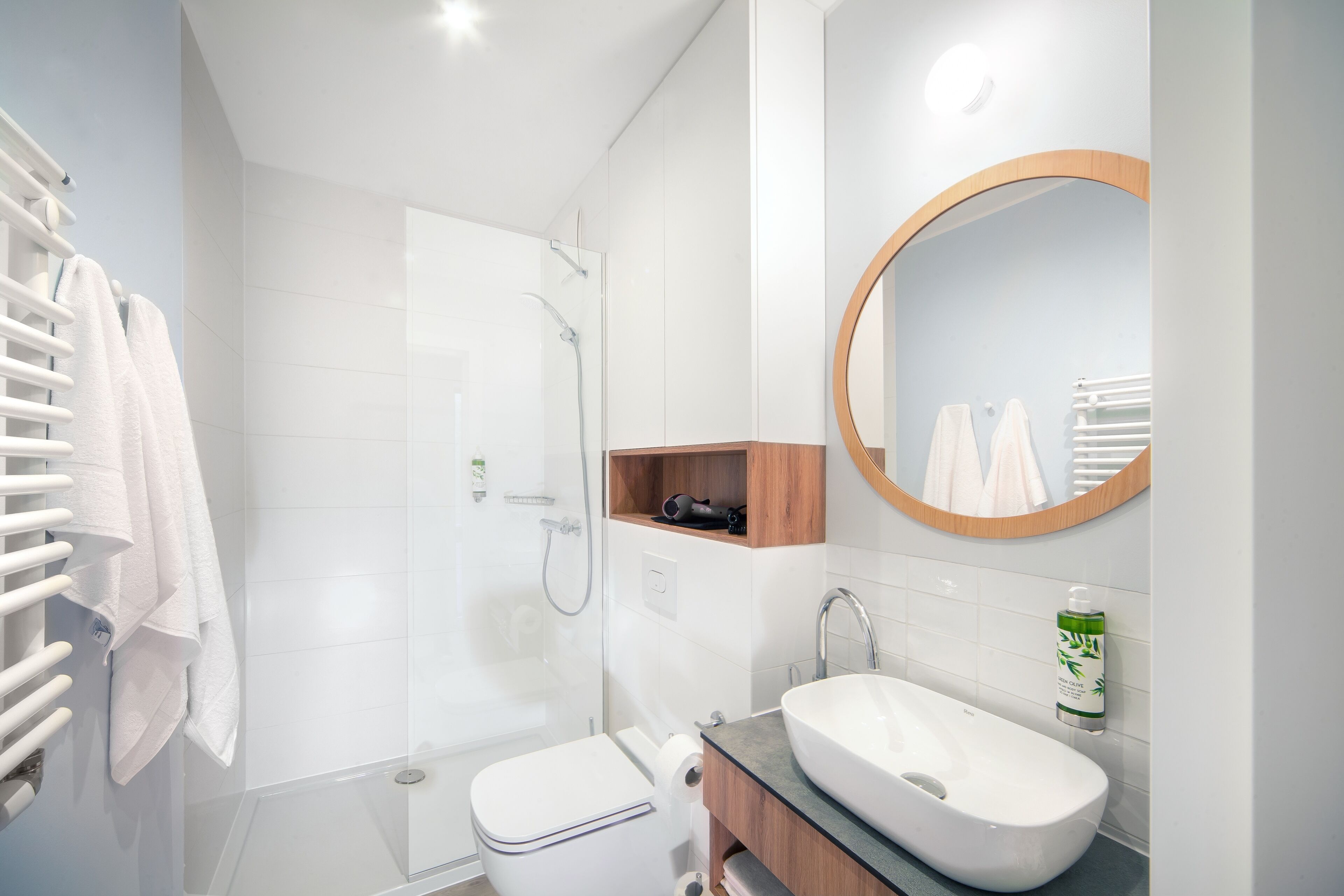 A modern bathroom with a glass-enclosed shower, a white toilet, a wooden vanity, and a round mirror.