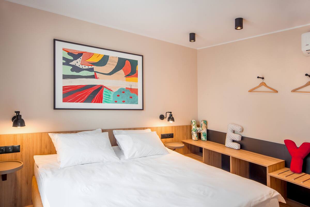 Ein modernes Hotelzimmer mit einem großen Bett, einem Nachttisch, einer Wandlampe, einem gerahmten abstrakten Gemälde und einem kleinen runden Tisch mit rotem Kissen.