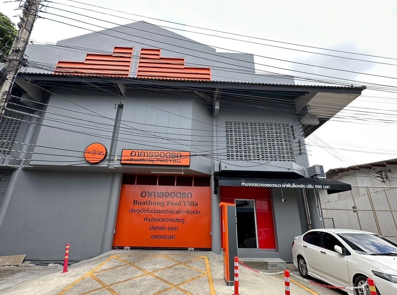 A building with a grey facade and an orange entrance. There is a sign for 'Buathong Pool Villa' and another sign for 'Buathong Pool Villa' in a different language. A white car is parked in front.