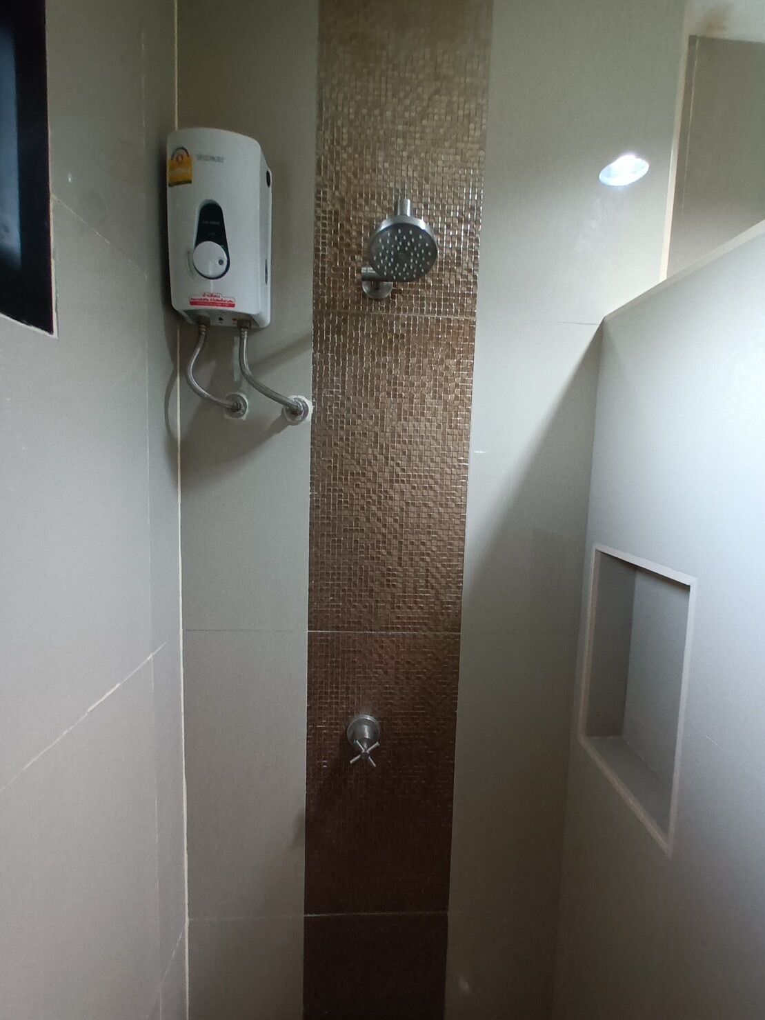 Un box de ducha estrecho con pared de azulejos, un cabezal de ducha manual y un calentador de agua instalado en la pared.