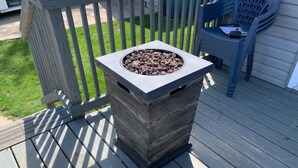 A square outdoor fire pit with a metal top and a wooden base, placed on a wooden deck. - Rye Beach Retreat · Cozy Cottage on Rye Beach with Lake Erie view! (Huron)
