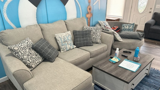 A living room with a grey sectional sofa, patterned cushions, a glass coffee table, and a blue rug. - Rye Beach Retreat · Cozy Cottage on Rye Beach with Lake Erie view! (Huron)