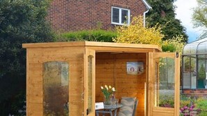 A wooden garden shed with a table and chair inside, a window, and a door. - CMK Bar & Spa (Milton Keynes)