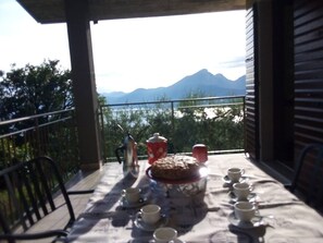 Un balcón con una mesa lista para el té, una tetera, tazas y un pastel, con vista a un cuerpo de agua y montañas.