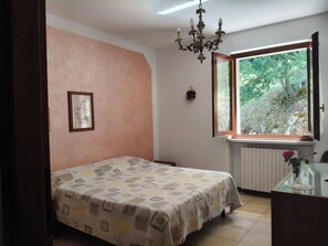A bedroom with a bed, a window with a view of greenery, a chandelier, and a framed picture on the wall.