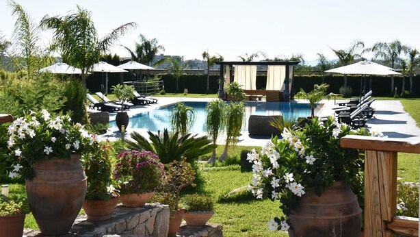 A pool area with lounge chairs and umbrellas, surrounded by potted plants and a grassy lawn. - VILLA FENICE (Sicilia)