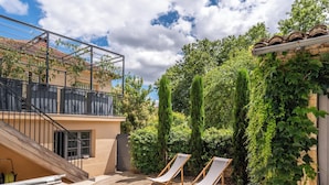 A poolside area with two lounge chairs, a wooden deck, and a view of a building with a tiled roof and greenery. - La Mignonne - Village house with swimming pool (Saint-Didier-au-Mont-d'Or)