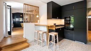 A modern kitchen with black cabinets, a wooden island with stools, and a visible oven. - La Mignonne - Village house with swimming pool (Saint-Didier-au-Mont-d'Or)