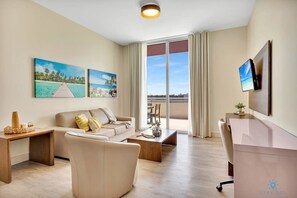 A hotel room with a sofa, coffee table, desk, and a large window with curtains. There are two paintings on the wall and a television mounted on the wall.