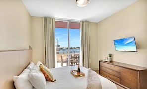 Quarto de hotel com cama, penteadeira de madeira, televisão e varanda com vista para o mar e um prédio distante.