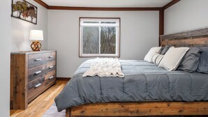 Une chambre à coucher avec une tête de lit en bois, une couvre-lit grise, un plaid blanc et une commode en bois dotée de tiroirs.