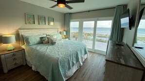 A bedroom with a bed, bedside tables, a ceiling fan, a TV, and a view of the beach through large windows. - Your Dream Getaway - A Slice of Heaven (St Joe Beach)