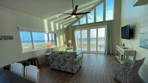A spacious living area with a large window overlooking the ocean, a ceiling fan, a dining table with chairs, and a television mounted on the wall. - Your Dream Getaway - A Slice of Heaven (St Joe Beach)