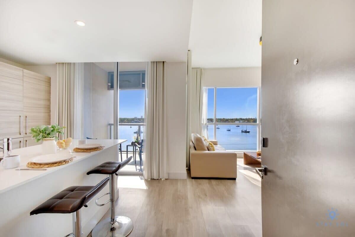 A modern kitchen with a large island, a dining area with bar stools, a sofa by a large window overlooking a body of water, and a view of boats.