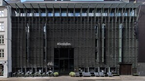 Une façade d’hôtel moderne, caractérisée par une structure extérieure en forme de grille, une entrée centrale et des espaces de repos en plein air.