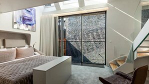 Une chambre moderne avec un grand lit, un bureau et une chaise. Une porte vitrée donne accès à un balcon offrant une vue sur la ville.