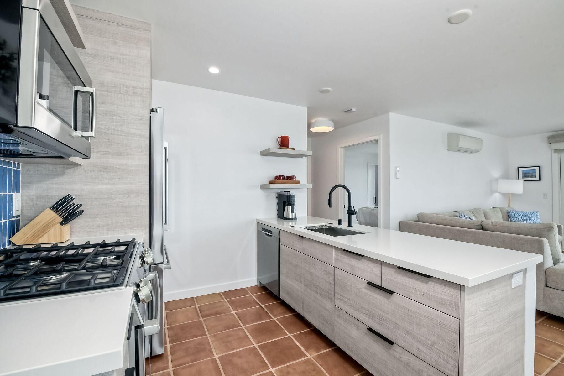 A modern kitchen with a gas stove, dishwasher, and white cabinetry. There is a dining area with a sofa and a coffee machine.
