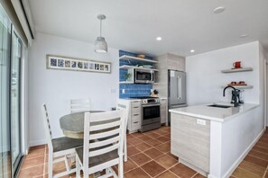 Cocina moderna con armarios blancos, horno empotrado, microondas y refrigerador de acero inoxidable. Hay una zona de comedor con mesa y sillas, y una lámpara colgante sobre la encimera.