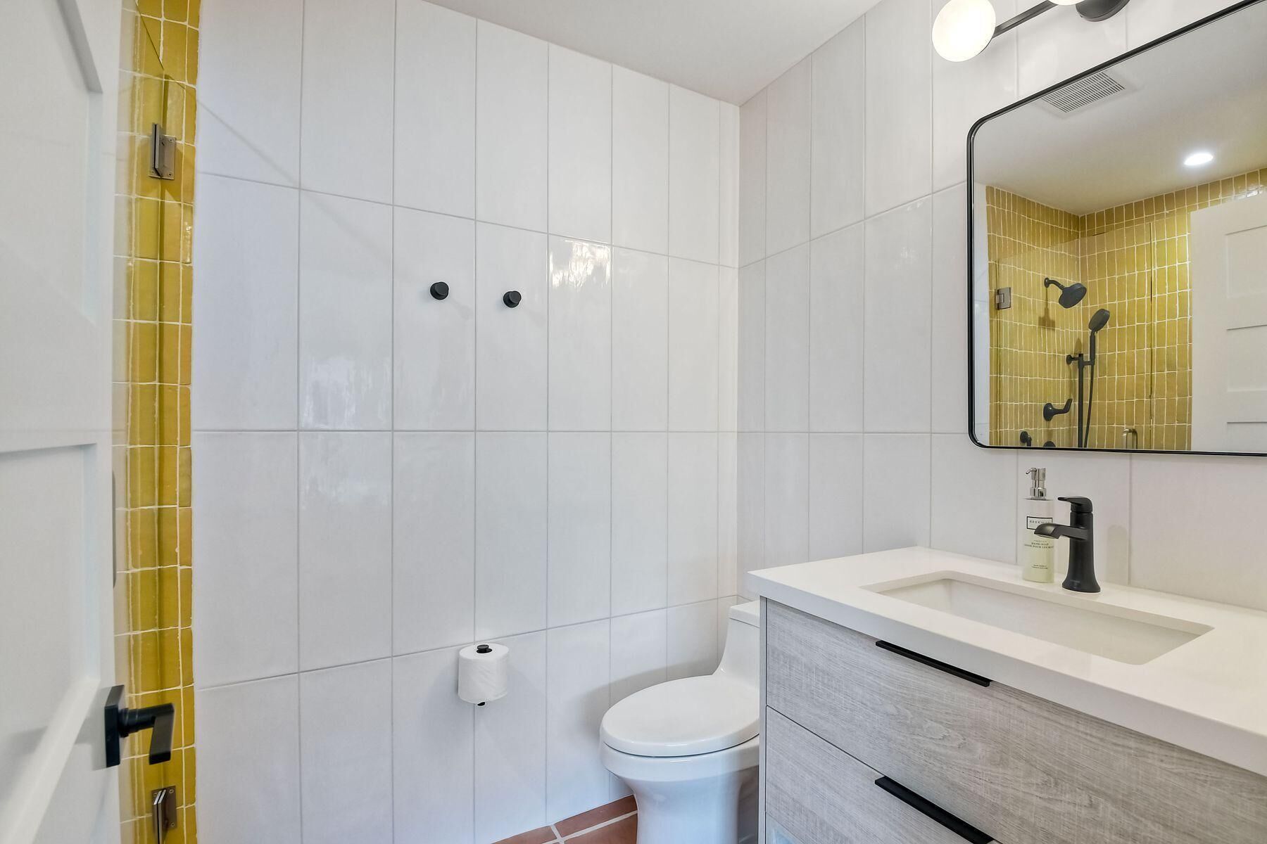 A modern bathroom with white tiles, a yellow-tiled shower area, a toilet, and a vanity with a mirror.