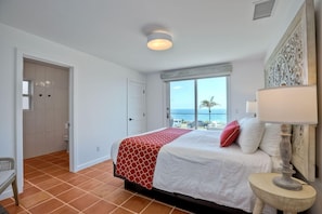 Um quarto com uma cama, uma janela com vista para o mar, uma porta e um abajur.
