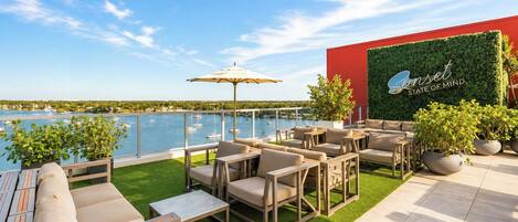 A terrace with outdoor seating, a view of the water, and a building with a green wall and signage.
