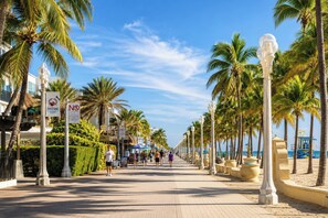 A beachfront promenade lined with palm trees and street lamps, with people walking and beach umbrellas in the distance.