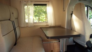 A compact RV interior with a dining table, cushioned seating, and a window with curtains. - Wohnmobil PLA Siena 385 (Krostitz OT Kupsal)