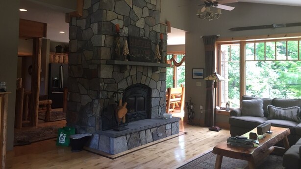 A stone fireplace with a mounted deer head, a ceiling fan, and a large window with a view of greenery. - Work Remote in Paradise on North Creek (North Creek)