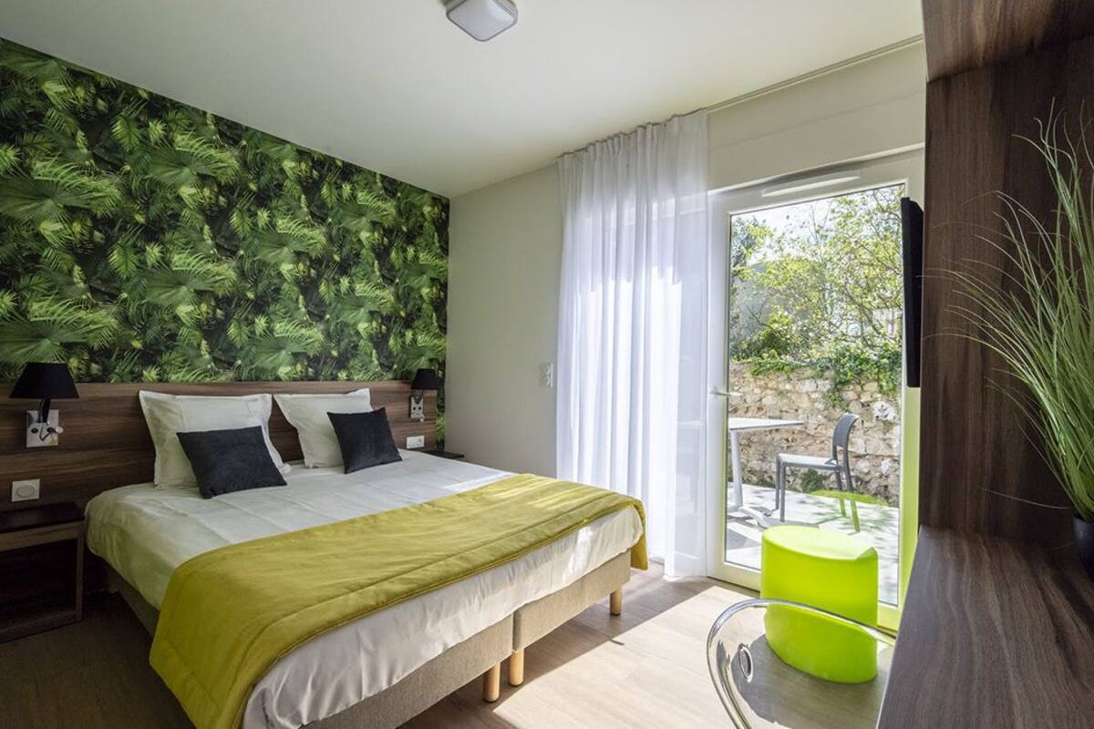 Une chambre à coucher avec un lit, une table de chevet, une lampe, un fauteuil vert et une vue sur l’extérieur.