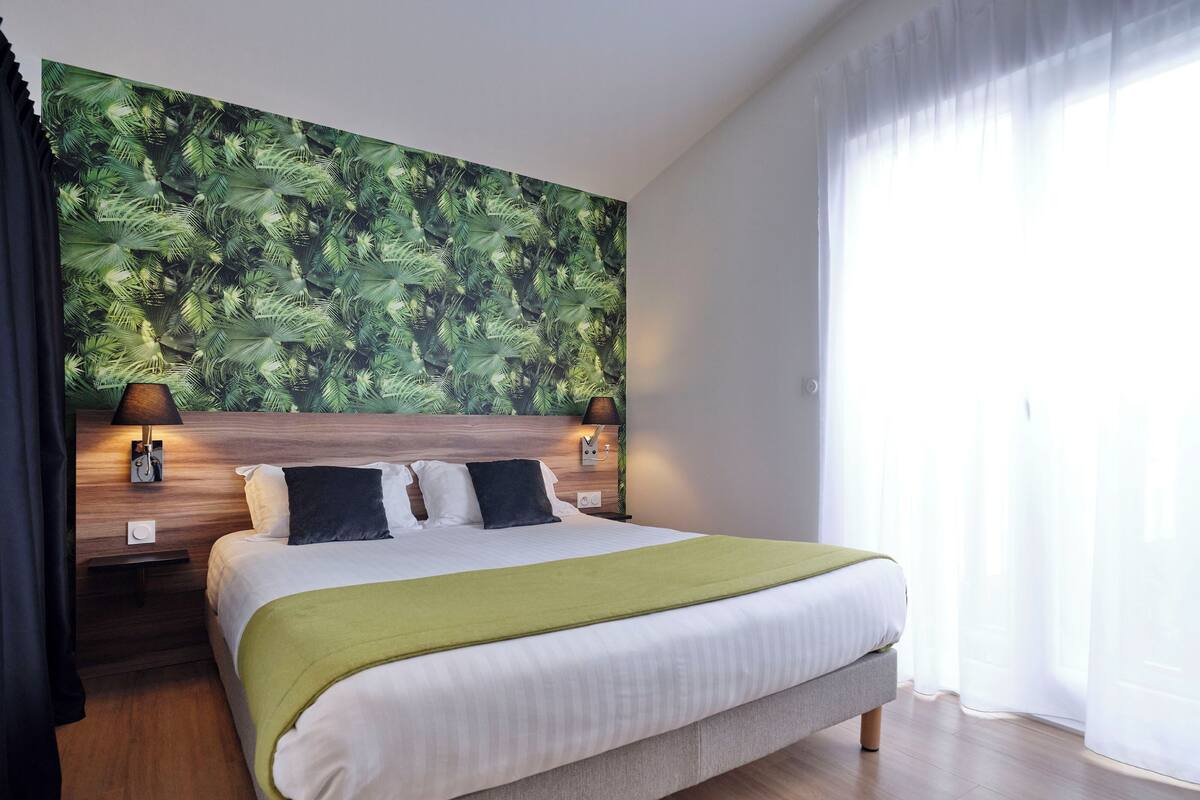 Une chambre d’hôtel avec un grand lit, une tête de lit ornée d’un motif de feuilles, deux lampes de chevet et une fenêtre avec des rideaux légers.