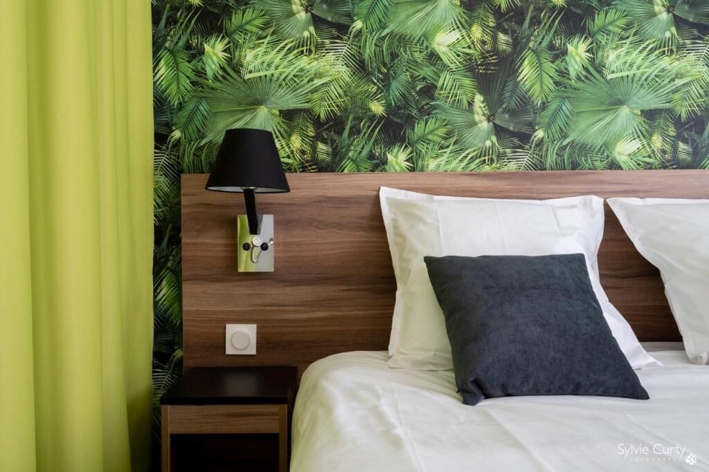 Ein Hotelzimmer mit Holz-Kopfteil, einem Nachttisch mit Lampe und einem Bett mit weißen und dunkelgrauen Kissen.