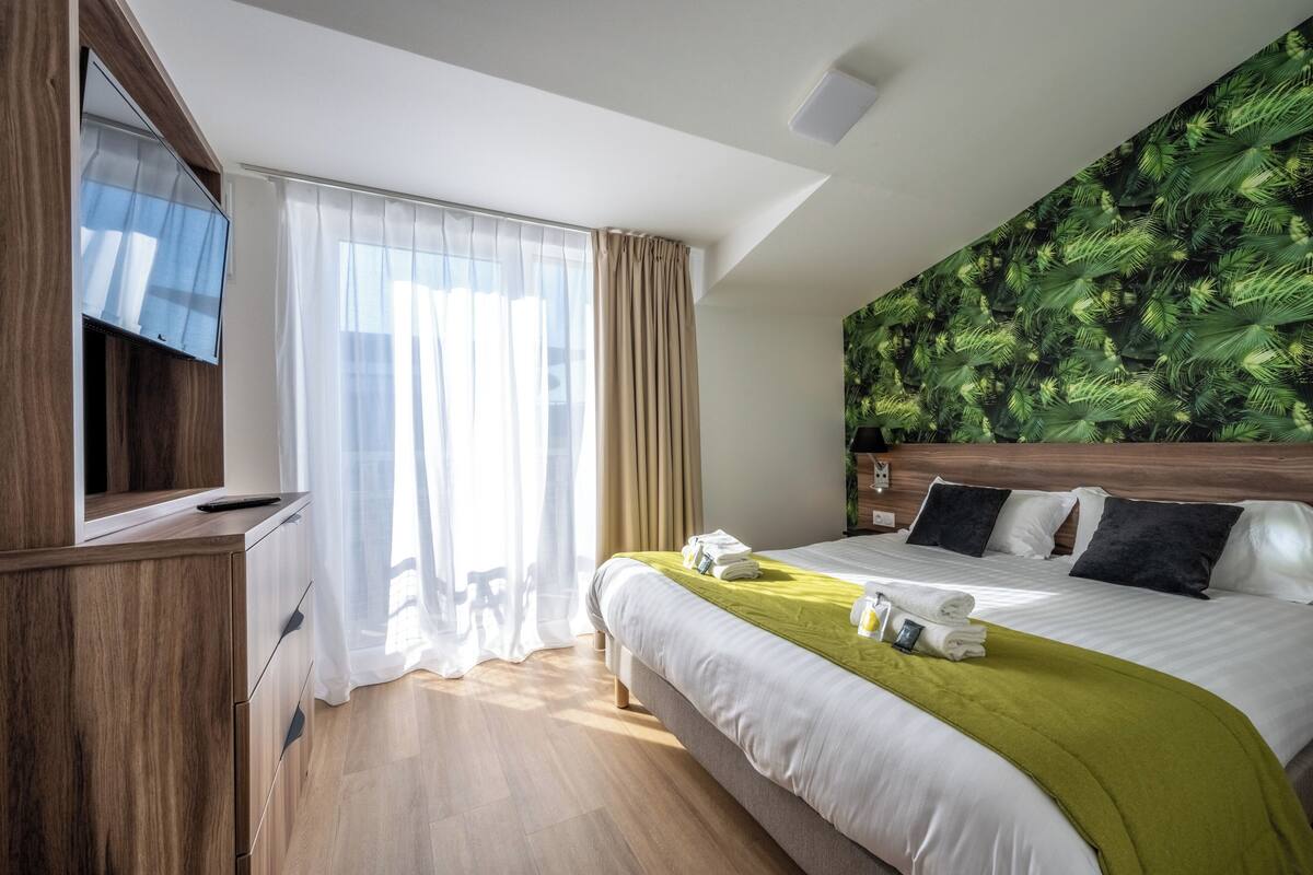 Une chambre d’hôtel avec un lit, un téléviseur fixé à un meuble en bois, une grande fenêtre avec des rideaux légers et un mur décoré d’un motif de feuilles vertes.