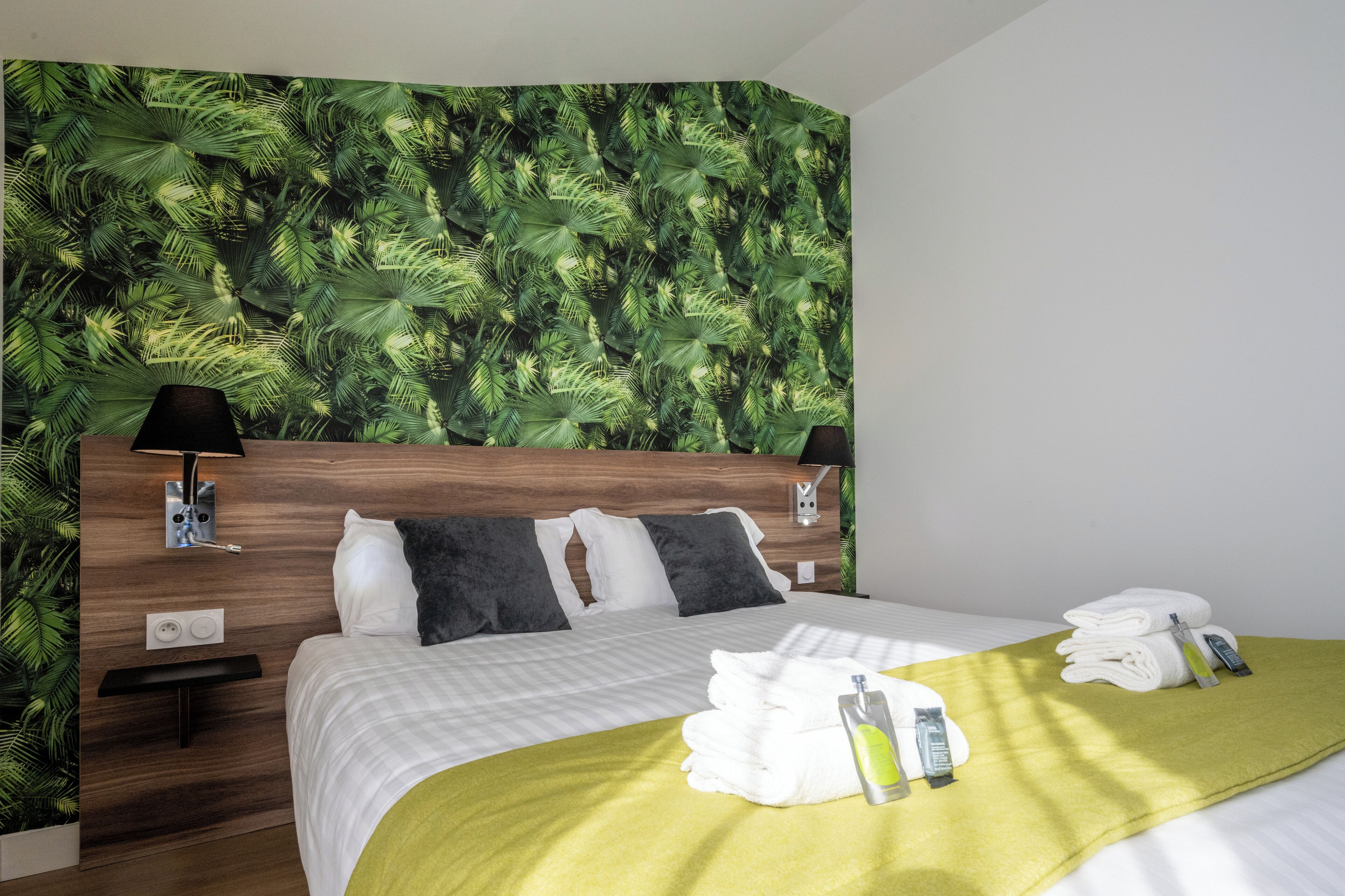 Habitación de hotel con cama, mesita de noche, lámpara de pared y pared verde con diseño de hojas.