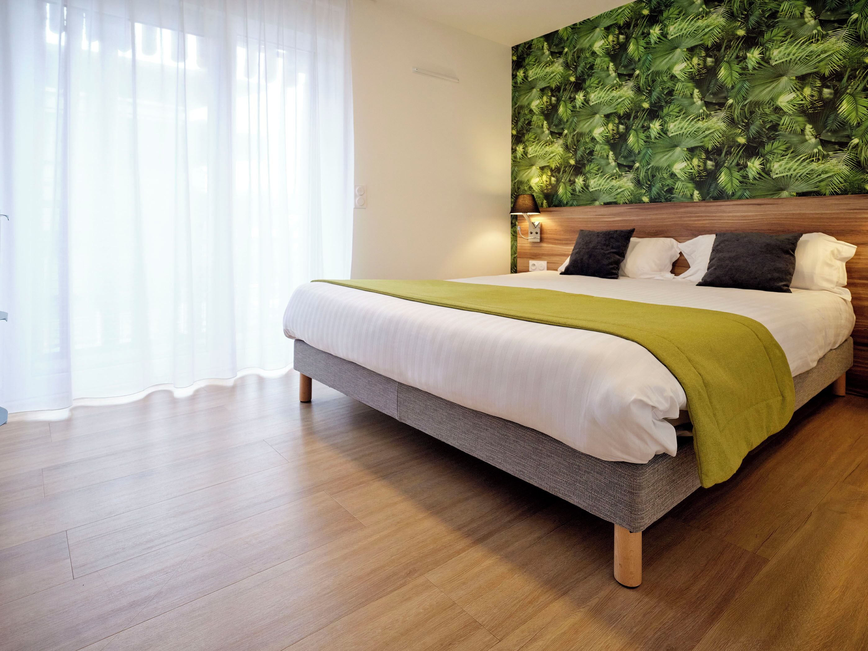 A modern hotel room with a large bed, wooden floor, and a tropical leaf-patterned wall.