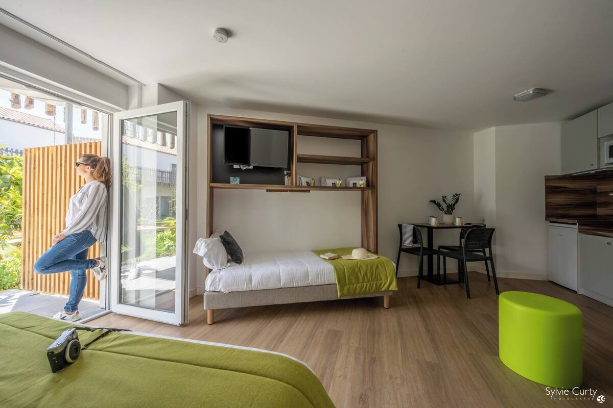 Une chambre moderne équipée d’un lit, d’un petit bureau, d’une télévision et d’un pouf vert.