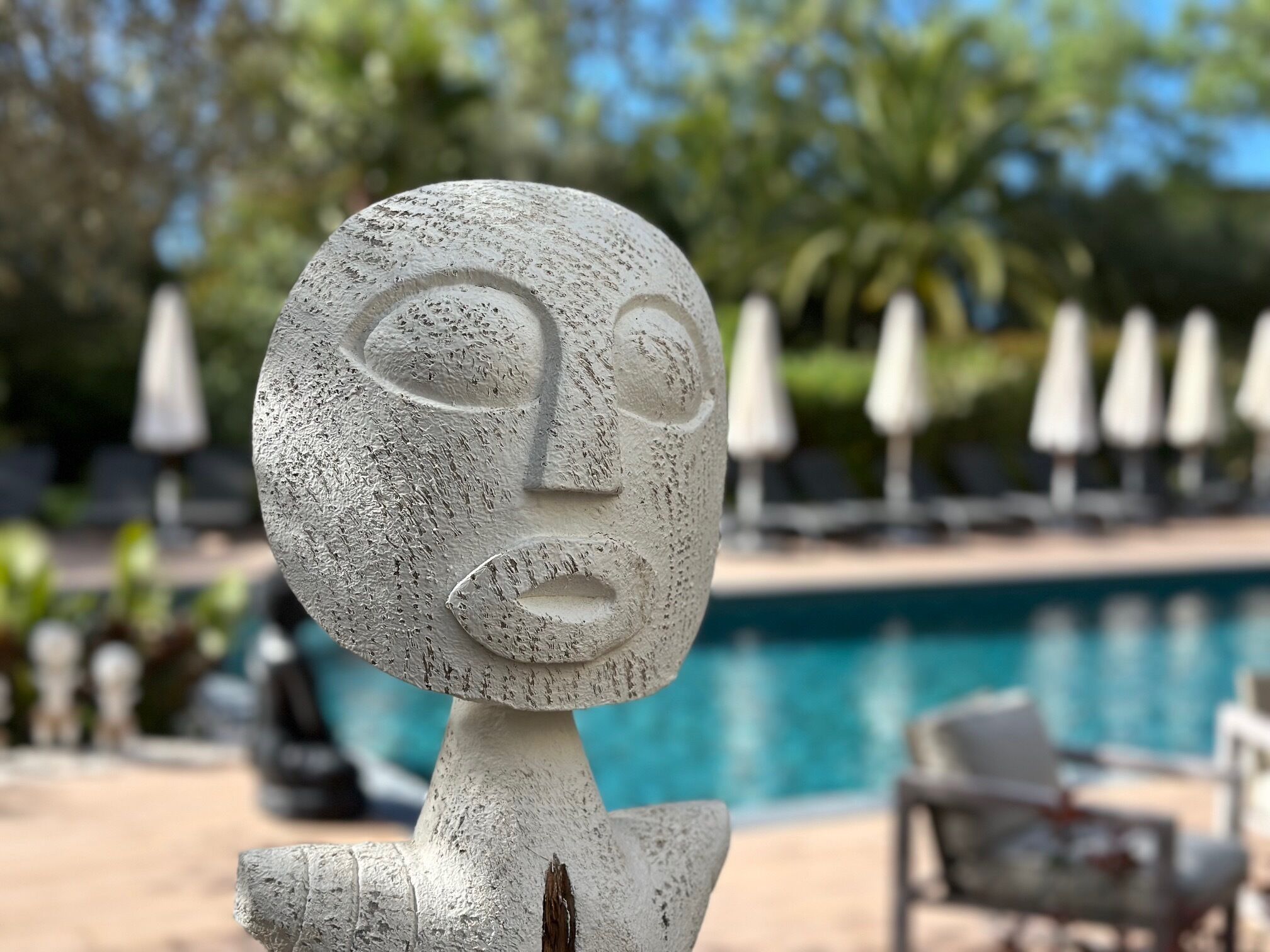 A sculpture of a face with a textured surface, positioned in front of a swimming pool with lounge chairs and umbrellas in the background.