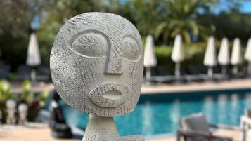 A sculpture of a face with a textured surface, positioned in front of a swimming pool with lounge chairs and umbrellas in the background.