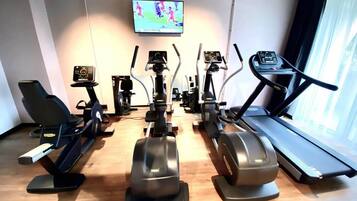 Un gimnasio con diferentes equipos de ejercicio, un televisor mostrando un evento deportivo y una ventana con cortinas.