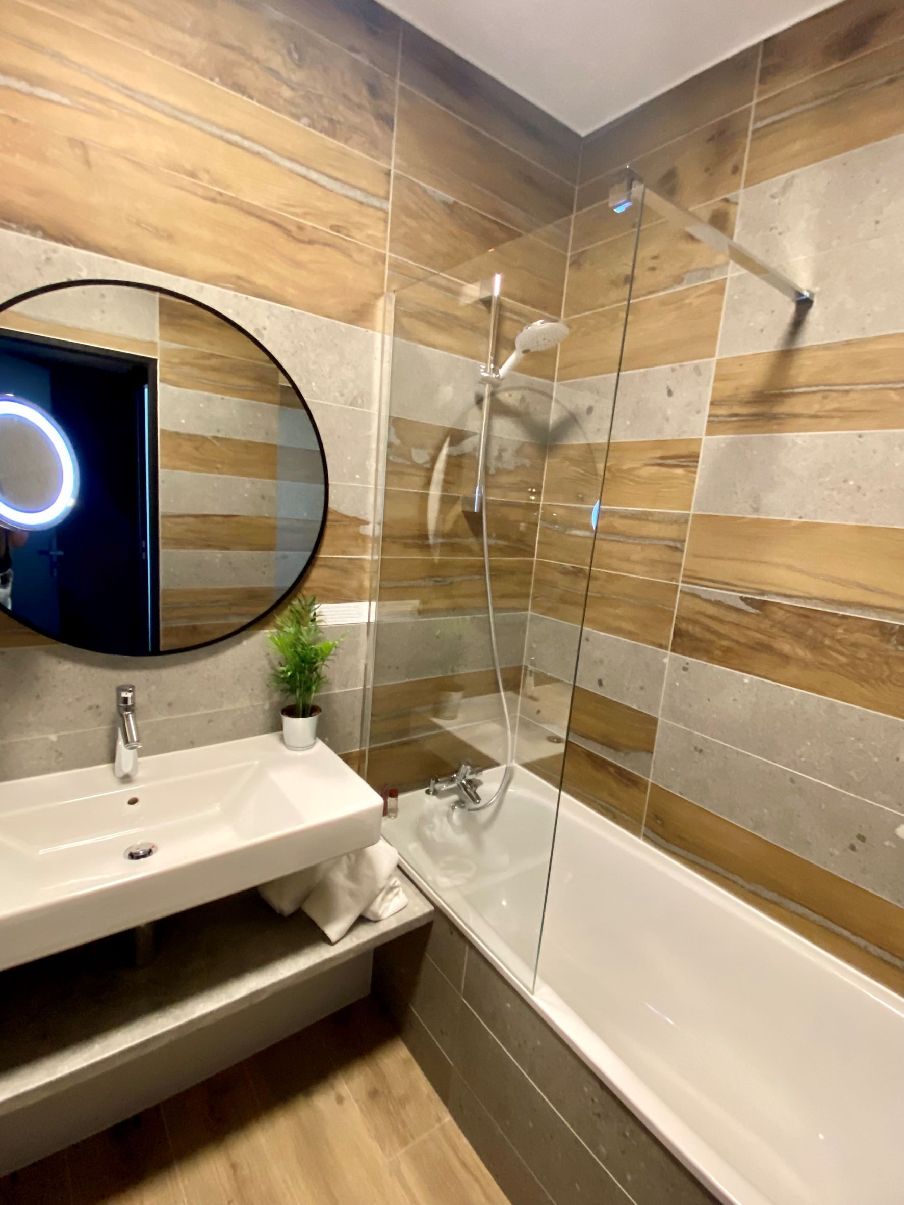 A modern bathroom with a glass-enclosed shower, a round mirror with a light, and a wooden wall design.