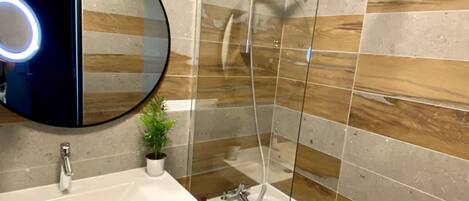 A modern bathroom with a glass-enclosed shower, a round mirror with a light, and a wooden wall design.