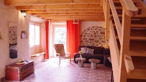 A cozy living room with a stone wall, wooden beams, a sofa, a chair, a small table, and a suitcase.