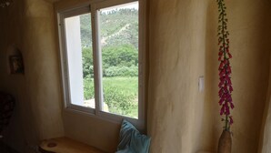 A room with a window overlooking a hillside, a wooden bench with a blue cushion, and a hanging flower arrangement.