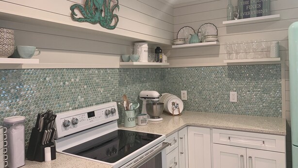 Uma cozinha com fogão branco, armários brancos e um painel de azulejos com mosaicos verdes. Há prateleiras com copos e uma peça de arte decorativa de polvo na parede.