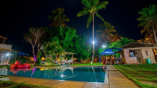 Ein Poolbereich bei Nacht mit freier Sicht auf den Pool, umliegende Palmen und ein kleines Gebäude mit einem blauen Sonnenschirm.