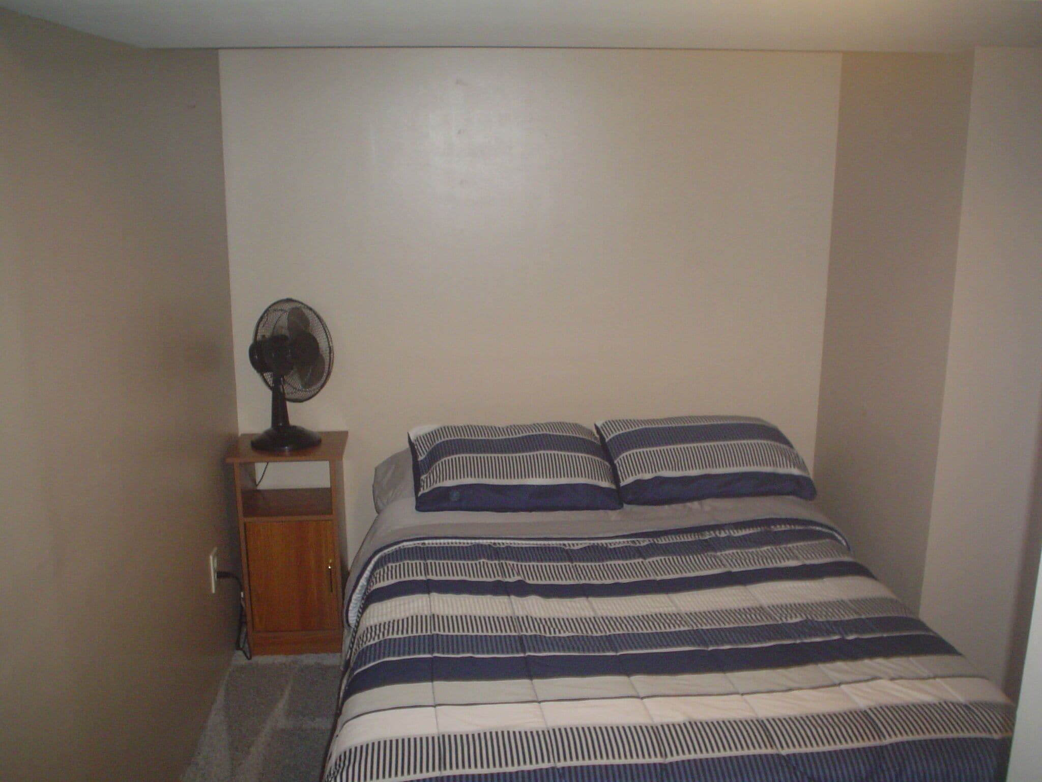 A small, single-bed room with a wooden nightstand, a standing fan, and striped bedding.