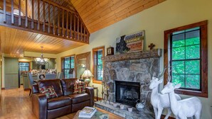 A cozy living room with a stone fireplace, a leather sofa, and a dining area visible in the background. - Red Top Treehouse Cabin in Mirror Lake and minutes to downtown-Dog Friendly! (Highlands)