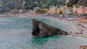 A coastal town with a prominent rock formation in the water, colorful buildings, and a beach with umbrellas. - Sciacchetrà Ground Floor Apartment with AC (Monterosso al Mare)