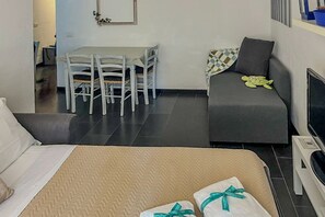 Hotellihuone, jossa on sänky, harmaa sohva, ruokapöytä tuoleineen ja televisio.